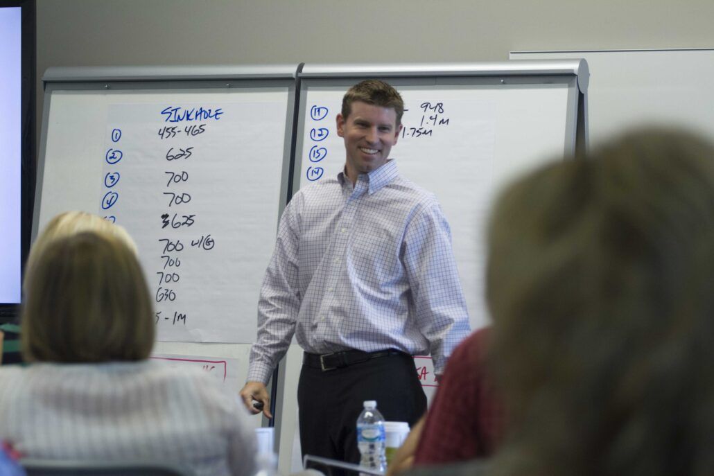 A man in a button-up shirt stands and smiles in front of two flip charts filled with handwritten numbers and notes.