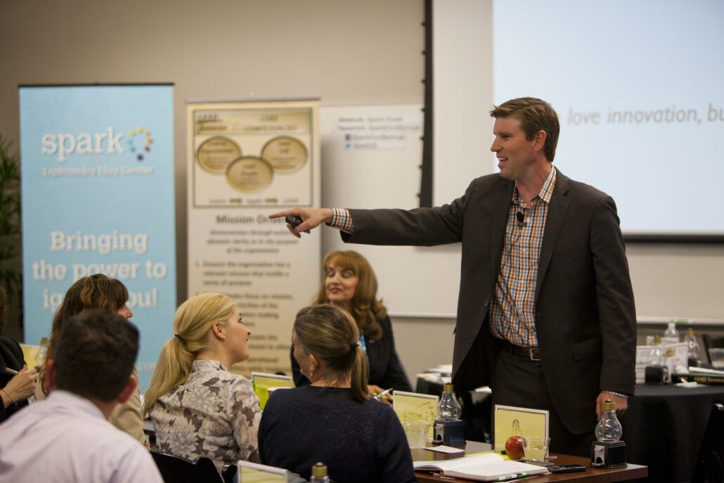 A man in a suit stands and gestures while speaking to a group of seated people in a conference room.