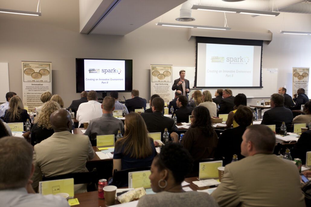 A speaker stands at the front of a conference room addressing a seated audience.