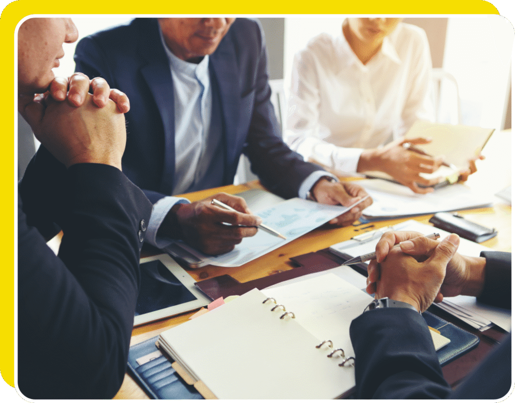 Four people sit around a table in a business meeting, with documents, a notebook, electronic devices, and charts spread out in front of them. One person points at a paper with a pen as others listen and learn how to negotiate effectively.