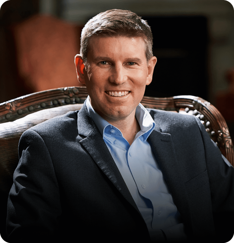 A man with short light brown hair, wearing a blue dress shirt and dark blazer, sits on a patterned chair and smiles at the camera against a dimly lit background—ready to help you learn how to negotiate with confidence.