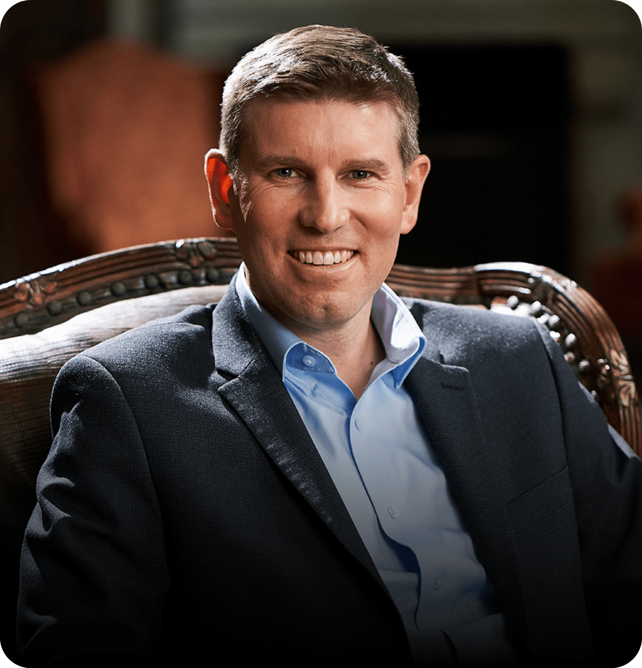 A man with short light brown hair, wearing a blue dress shirt and dark blazer, sits on a patterned chair and smiles at the camera against a dimly lit background—ready to help you learn how to negotiate with confidence.